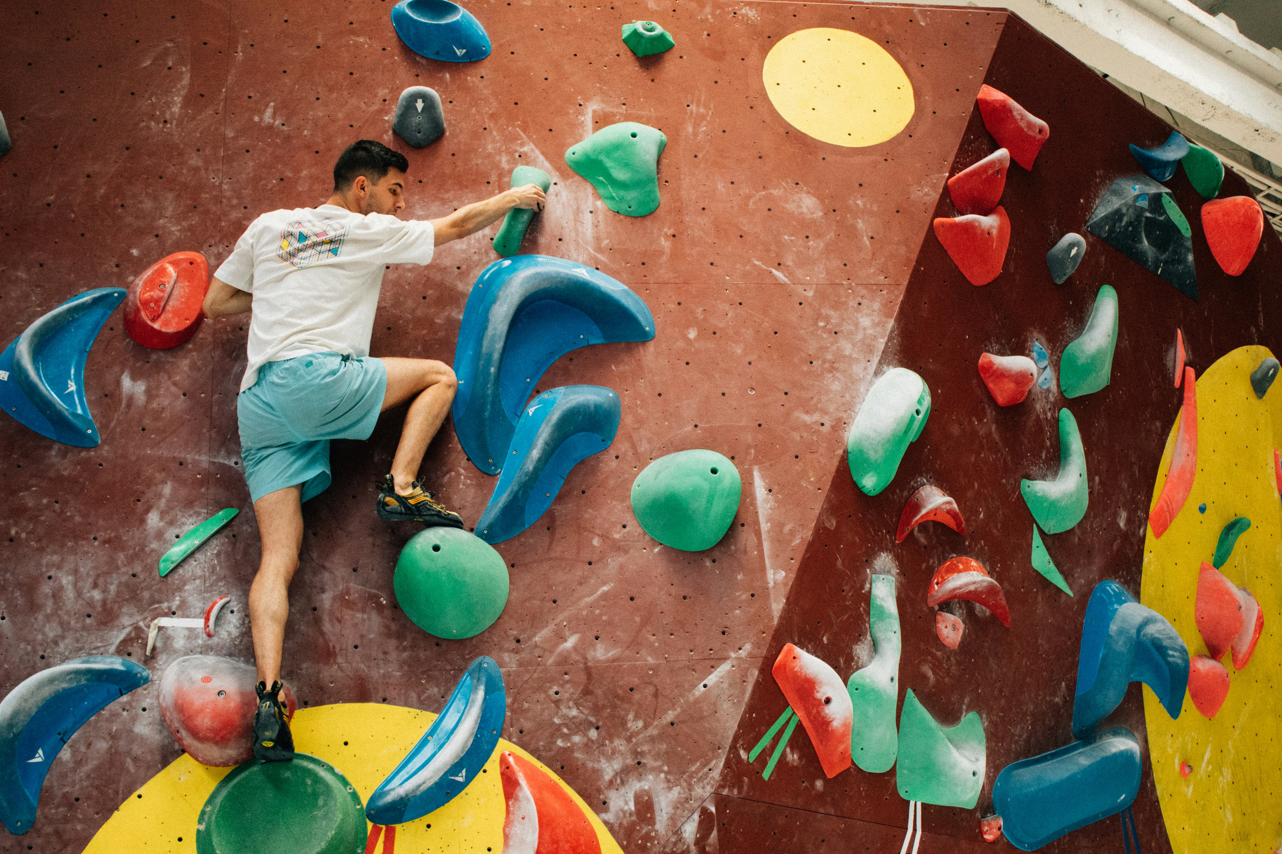Climber on an indoor bouldering wall with large green, blue, red, and yellow holds